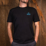 A man wearing a black short-sleeve t-shirt with a small blue 'Vice Versa' logo on the left chest, standing against a rustic wooden plank wall.